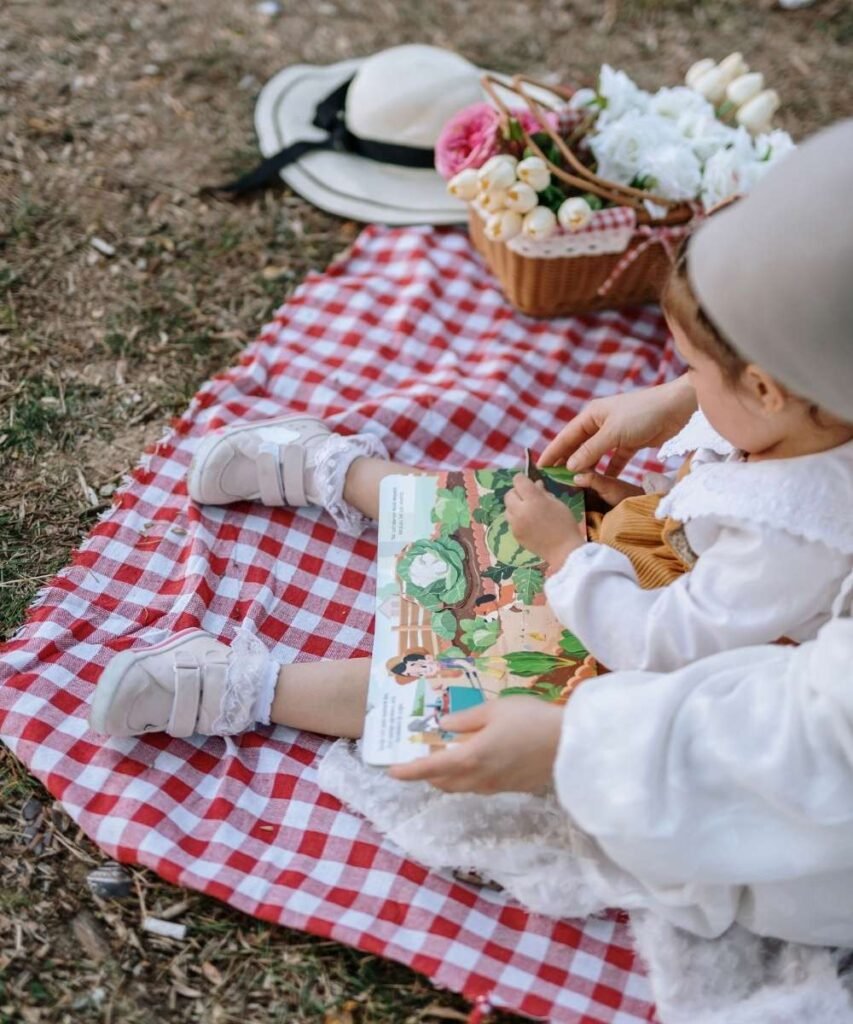 A young toddler wearing a white outfit sitting outdoors on a checkered picnic blanket reading a picture book.
