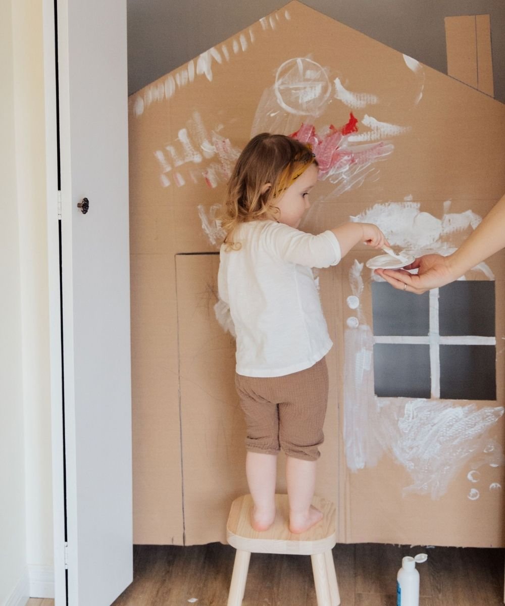Toddler standing indoors coloring with a crayon on a large cardboard box flattened on the wall