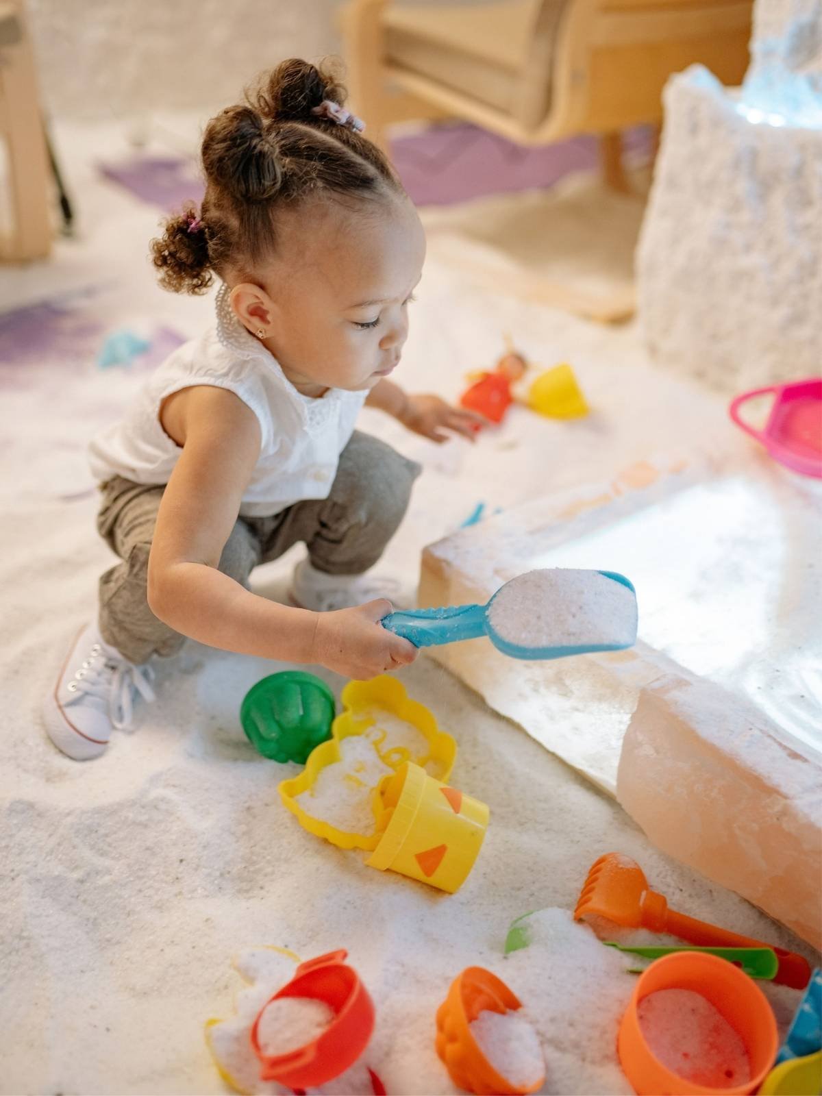 Alt Text: Toddler crouching on floor scooping white sensory material with a blue plastic shovel