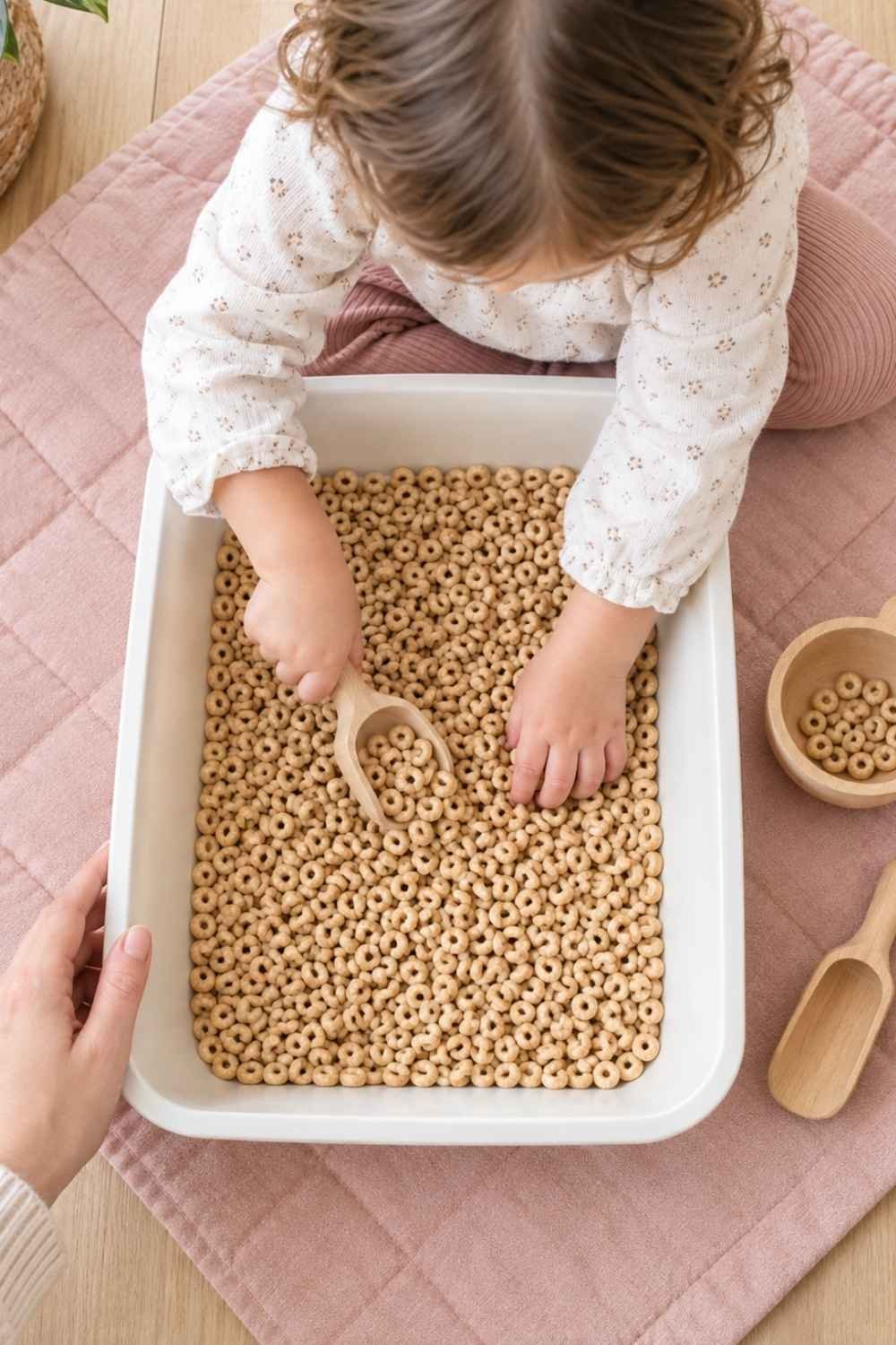 Young toddler playing with a safe dry cereal sensory bin on a low tray with wooden scoops.