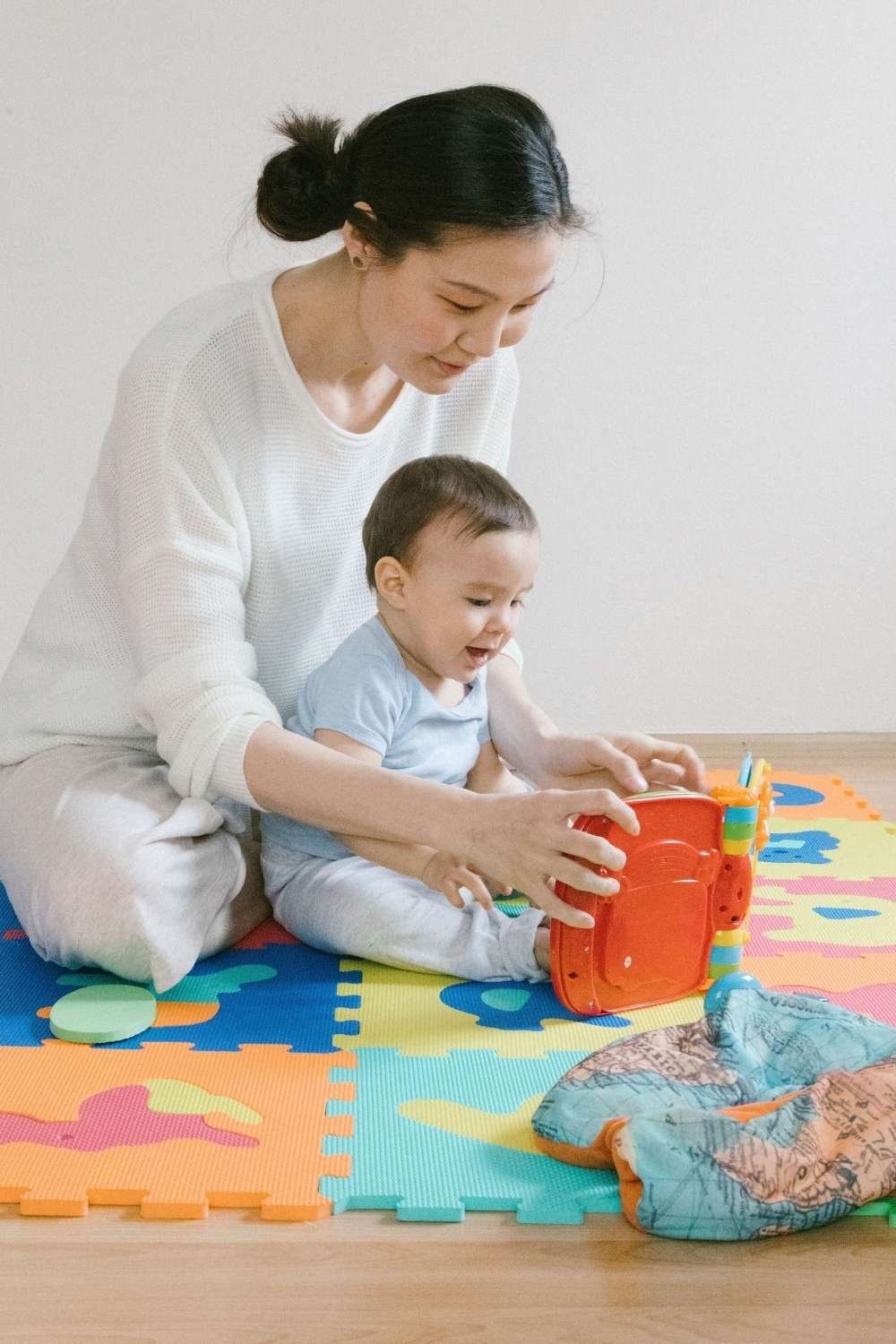 A mother and toddler boy sitting on the floor together engaging in safe sensory bin play with colorful toys and containers.